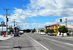 A small-town straight street, flanked by shops, parked cars and the occasional small tree. A Caltex petrol station is on the left, a two-storey pub painted bright blue and yellow is on the right.