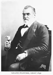 Elderly Caucasian male, wearing a formal suit, with a watch fob, sitting in a chair and holding a cane.