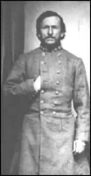 Black and white photo shows a man from his head to his knees, wearing a long gray military coat. His right hand is thrust under the coat, Napoleon-style.