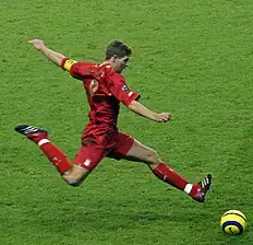 Steven Gerrard wearing a football kit, about to strike a ball. On his forearm, he is wearing an armband; the letter "C" to represent "Captain" is visible.