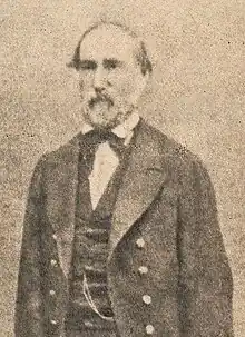 A black a white headshot of a man with a beard wearing a suit.