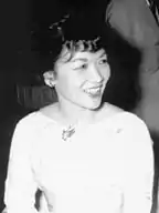 A middle-aged lady wearing a light-colored dress and with short hair, fluffy at the front, sits at a dinner table smiling.