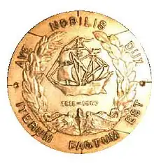 A round metal object features a sailing ship in the center. In a circle around the edges are the Latin words "Ave Nobilis Dux, Iterum Factum Est".