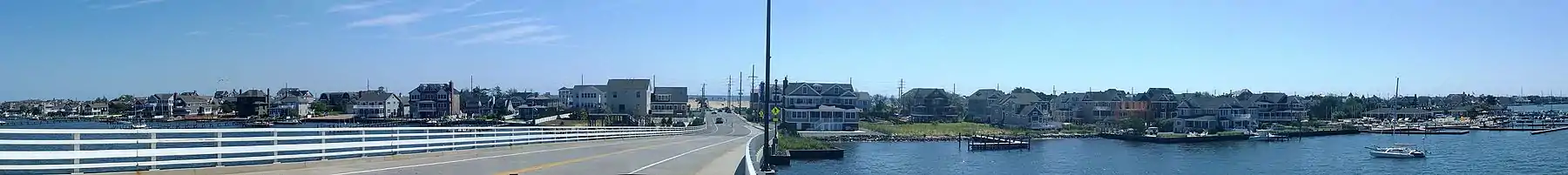 Panorama of town showing houses along the length of the image. A road descends from a bridge in the center to ground before terminating at a signalized intersection. An ocean is located in the background in the center of the image.