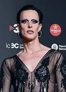 a photo of Marina from the chest up. She is wearing a black mesh shirt, embellished with sequins and crystals. Her hair is slicked back and she is wearing hoop earrings.