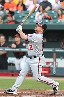 A stocky bearded man wearing a gray baseball uniform with red and navy-blue trim watches after following through on a swing