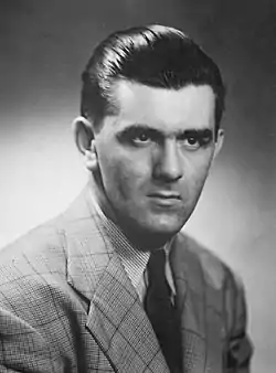 Upper body of a young man in a suit and tie with slicked back hair and a serious look on his face