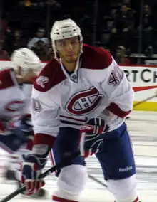 A Caucasian ice hockey player in his mid-twenties. He holds his hockey stick in a ready position while looking forward. He wears a white, visored helmet and a white jersey with red trim.