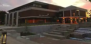 A three-floor brown building of brick and concrete is pictured with a walkway and plaza in front of it and water feature in the foreground, with the middle of the building featuring a large reading room which overlooks the plaza. The building is topped by a green slanted metal roof. To the left of the picture, four brass panels featuring reliefs of literary themes, concepts, characters and writers are contained within four concrete structures lit by LED lighting