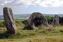 two weathered stones standing at an angle on a grassy hill, with a third doughnut-shaped stone between them