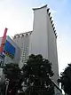 Ground-level view of two concrete towers; the one on the right is more prominent, and has a windowless lateral facade with triangular structures protruding from the roof.