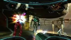 View of a futuristic looking room; two enemies wearing powered armor are approaching the player, and one is being hit by the player's weapon (a large cannon), which is visible in the corner of the screen. The image is a simulation of the heads-up display of a combat suit's helmet, with a crosshair surrounding the enemy and two-dimensional icons relaying game information around the edge of the frame.