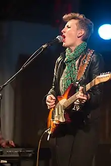 A 32-year-old woman is show in left profile. She is singing into her microphone while strumming an electric guitar.