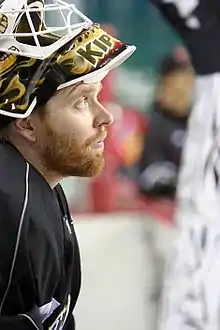 Kiprusoff in the white Flames away jersey with a stylized red C logo on his chest and his mask raised above his forehead stares down at the ice as he is skating.