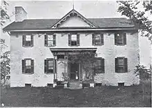 A two-story building made of light brick, featuring several windows with black shutters, a small front porch, a single gable in the middle of the roof, and a chimney on the left