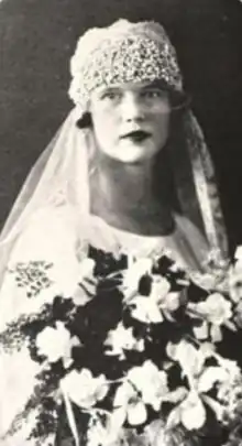 A young white woman in a wedding dress and veil, with a close-fitting textured cap fit over her brow; she is holding a large bouquet of light-colored flowers