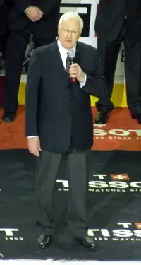 Costello in a suit and tie speaking into a microphone at an on-ice presentation