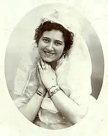 A young white woman with dark curls, smiling, hands together under her chin; she is wearing light-colored lacy clothes including a headpiece, and several bracelets