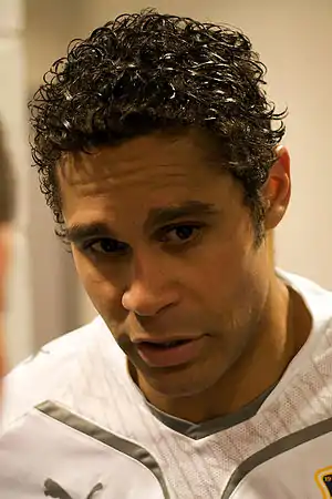 A close-up photograph of a man with brown-black hair wearing a white training sweater.