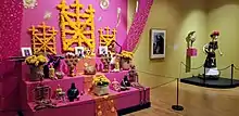 Photo of museum exhibits. At left, a brightly colored three-tiered altar featuring reminders of lost loved ones like food, sculptures, and photographs. At right, a skeleton wears a dark dress and bright flower hat.