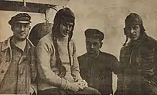 Photograph of a women wearing a hat with ear-flaps over long braids between one man on the left and two men on the right