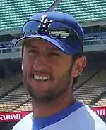 a smiling man with a LA Baseball cap and sunglasses above the caps visor smiles while wearing a stubble beard.