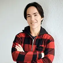 Head and shoulders image of a woman with short-straight black hair, wearing a hooded red and black checked jacket, with her arms crossed in front of her chest