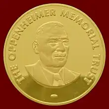 A golden medallion with an embossed image of a mustached man. Surrounding the bust in bold uppercase type is embossed the words, THE OPPENHEIMER MEMORIAL TRUST.