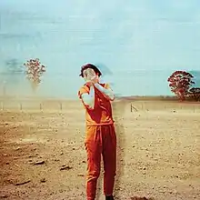 A woman stands in the middle of a field, covering her face with her hands