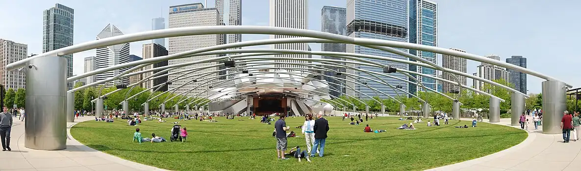 Futuristic view of a green lawn with people scattered on it, beneath a metal trellis supported on shiny metal pillars. The trellis leads to a bandshell surrounded by curved plates of shiny metal, with many tall skyscrapers in the background.