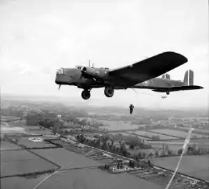 Man hangs suspended from aircraft flying over tree lined fields