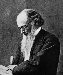 Head and shoulders photograph of a man in an overcoat.  He has a full beard with sideburns, has a shaved upper lip and is balding.  He wears a serious expression and is looking down at a paper with an insignia drawn on it.