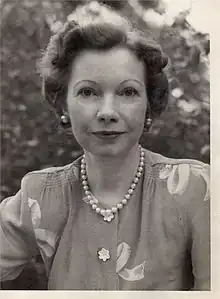 A white woman with dark hair in a set style, wearing a strand of beads or pearls and a print blouse, photographed outdoors