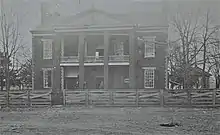 A sepia-tint black and white photograph of the front of an old two story brick building with a large porch and portico behind a wooden fence. A man stands in the doorway, a small painted sign advertising a lawyer's services is hung near the front door, and a gazebo is located to the right of the building.