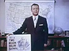 A still shot from the 1965 film Perversion for Profit showing a man in a suit holding a placard. The placard features a cartoonish image of a leering man and the phrase "It's a two billion dollar business"