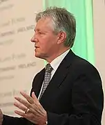 talking, clean-shaven man with longish salt-and-pepper hair wearing a dark suit and grey tie, speaking at a podium