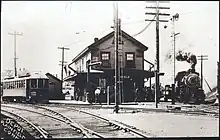 A two-story wooden railway station at a junction of two lines. A streetcar is on the left line, and a steam train on the right line.