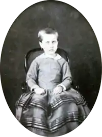 Photograph of a young Isabel with hair gathered at the back and wearing earrings seated on a small, cane back armchair