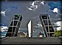 A stone monument stands in the center. Two skyscrapers leans to the center. The sky has been ominously altered.