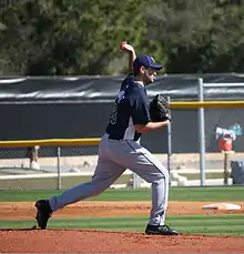 A man throwing a baseball wearing a navy-blue baseball jersey and cap and gray baseball pants