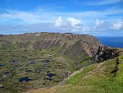 Colour photo from a cliff top down onto a pond strewn marsh backed by high cliffs, an area of sea is visible through a gap in the cliffs.