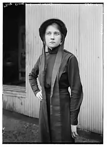 A young white woman, standing, wearing a dark bonnet-style hat with long fabric ties, and a plain dark dress with long sleeves and skirt