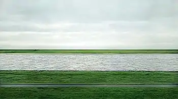 A photograph of the Lower Rhine river flowing horizontally through green fields under an overcast sky in Germany