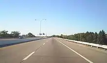 Ground-level view of two lanes of a divided freeway; the opposing lanes of traffic are separated by a narrow concrete barrier. A dense forest is visible to the left.