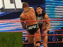 Two dark haired men standing in a wrestling ring with red ropes, one with his back to the camera. The man with his back to the camera is wearing short black wrestling tights with the name 'Orton' written on them in white lettering, and has a tribal design tattoo on his back. He is throwing a punch at the other man, who is facing the camera with a look of surprise, and is wearing short purple wrestling tights and a black top with a white design on it.