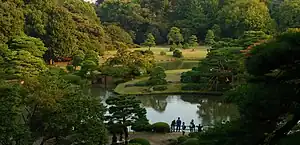 A pond in a wooded park with a bridge and grassland.