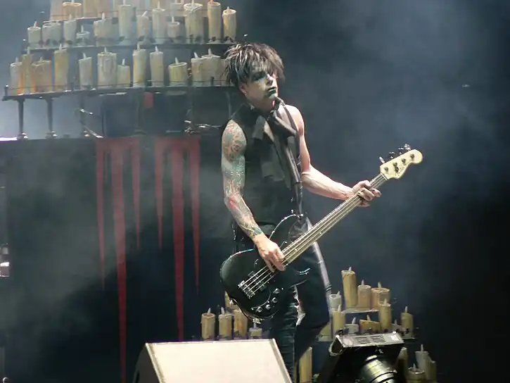 Rob Holliday wearing a black sleeveless goth outfit, standing onstage playing bass guitar