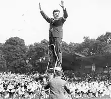 A man in a suit stands on a step ladder to conduct a large group of drum musicians