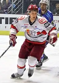 An ice hockey player skating towards the camera. He is wearing a red helmet and a red and white uniform.