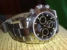 A wristwatch manufactured by the Swiss brand Rolex. The watch is constructed from stainless steel with a sapphire crystal covering a black dial. The watch has a timing feature which uses the large seconds hand which makes one complete revolution over a single minute, and includes three sub-dials. Two of these sub-dials track elapsed minutes and hours, and the other sub-dial tracks seconds for the current time, as the main seconds hand is used for the timing feature.